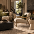 Load image into Gallery viewer, A modern living room scene featuring a cream upholstered accent chair with gold-toned metal arms and legs, styled with an olive green pillow. The chair is set on a textured beige rug next to a dark wood coffee table and a stone side table, with a matching cream sofa and indoor plants in the background.