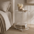 Load image into Gallery viewer, A clear acrylic side table with a rounded rectangular shape, shown in a styled bedroom next to a bed. The table has a top surface and a lower shelf holding books, with decorative items including a vase of white flowers and a candle on top.