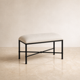 Load image into Gallery viewer, A rectangular upholstered bedroom bench with a cream fabric seat cushion and a black metal frame, set against a warm beige wall and light stone floor.