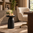 Load image into Gallery viewer, A modern black round side table with a textured, tapered pedestal base stands in a styled home office or living room setting next to a cream bouclรฉ armchair and a wooden desk.