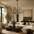 Load image into Gallery viewer, A modern black chandelier with gold-finished candle sleeves hangs in a styled living room with a cream sectional sofa, dark wood coffee table, and neutral-toned decor.