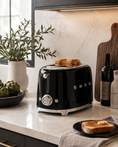 Load image into Gallery viewer, black toaster with silver accents sits on a white marble kitchen countertop, toasting two slices of bread. The scene is styled with a bottle of olive oil, a jar of salt, a wooden cutting board, and a plate of toast in a modern kitchen setting.