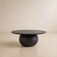 Load image into Gallery viewer, A round black stone coffee table with a textured top and a rounded pedestal base, set against a minimalist beige wall and floor.