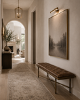 Load image into Gallery viewer, A large framed black and white landscape art print with a gold frame hangs on a beige wall in a hallway, above a brown bench with a gold metal frame. A patterned area rug runs along the floor, and an arched doorway in the background reveals a cozy living room with a fireplace and plants.