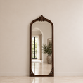Load image into Gallery viewer, An ornate full-length floor mirror with a dark carved wood frame, leaning against a neutral beige wall in a bright, modern interior.