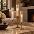 Load image into Gallery viewer, Three brass table candle holder with clear ribbed glass cylinders and lit candles, arranged on a marble coffee table in a cozy living room setting.