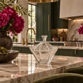 Load image into Gallery viewer, A clear faceted glass candy dish with a matching lid sits on a polished marble kitchen island. The background shows a luxury kitchen with dark green cabinets, gold fixtures, and a vase of pink peonies on the counter.