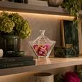 Load image into Gallery viewer, A clear glass lidded candy dish filled with pink dried flowers sits on a wooden shelf beside a green ceramic vase with hydrangeas, books, and framed decor in a styled interior setting.