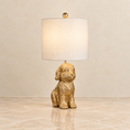 Load image into Gallery viewer, A table lamp with a textured gold dog-shaped base and a white fabric drum shade, shown on a polished marble surface against a neutral beige wall.