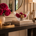 Load image into Gallery viewer, A clear crystal bowl filled with pink and gold decorative pom-pom ornaments sits on a stack of Louis Vuitton books on a dark wood console table. The scene is styled with a vase of deep pink peonies, a lit candle, and a table lamp in an elegant interior setting.