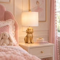 Load image into Gallery viewer, A gold dog-shaped table lamp with a white drum shade sits on a white nightstand in a pink bedroom setting next to a bed with a pink headboard and plush bunny.