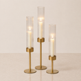 Load image into Gallery viewer, Three gold-toned candlestick holders with clear ribbed glass cylinders and white candles, arranged on a light beige surface against a neutral cream background.