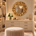 Load image into Gallery viewer, A luxury walk-in closet featuring a large round gold leaf mirror above a cream-colored double dresser with gold handles. In the foreground is a round tufted beige ottoman, with a vase of white flowers and candles on the dresser.