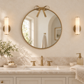 Load image into Gallery viewer, gold round mirror with a gold-toned frame and bow detail at the top, mounted above a white vanity with a gold faucet. The scene is styled like a bathroom with two matching wall sconces, marble countertop, soap dish, and decorative plants