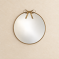 Load image into Gallery viewer, gold roundwall mirror with a distressed brass frame and a matching bow detail at the top, mounted on a textured beige wall