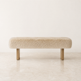 Load image into Gallery viewer, A modern rectangular bench with a thick, textured cream-colored sheepskin or faux fur cushion and light natural wood legs, set against a minimalist off-white wall and light marble floor.