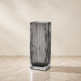 Load image into Gallery viewer, A tall, rectangular ribbed glass vase in a smoky translucent gray color, standing on a beige marble surface against a textured beige wall.