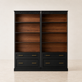 Load image into Gallery viewer, A tall black bookcase with two open wooden shelves on each side and four drawers at the bottom, accented with gold handles, shown against a neutral beige background.