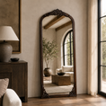 Load image into Gallery viewer, wall mirror with ornate dark carved wood trim stands in a styled room with beige walls, a wooden cabinet, a lamp, and a window looking out onto greenery.