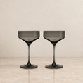 Load image into Gallery viewer, Two dark gray smoked glass coupe glasses on a light beige marble surface against a textured beige background.