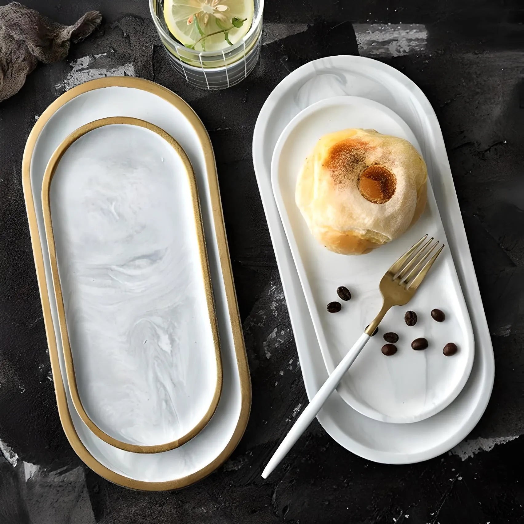 Two House of Effy MINIMALIST OVAL SERVING PLATES: the left one is empty with golden edges, while the right complements a sophisticated tableware setting with a pastry, fork, and coffee beans. An elegant drink garnished with a flower and lemon slice sits adjacent to the trays.