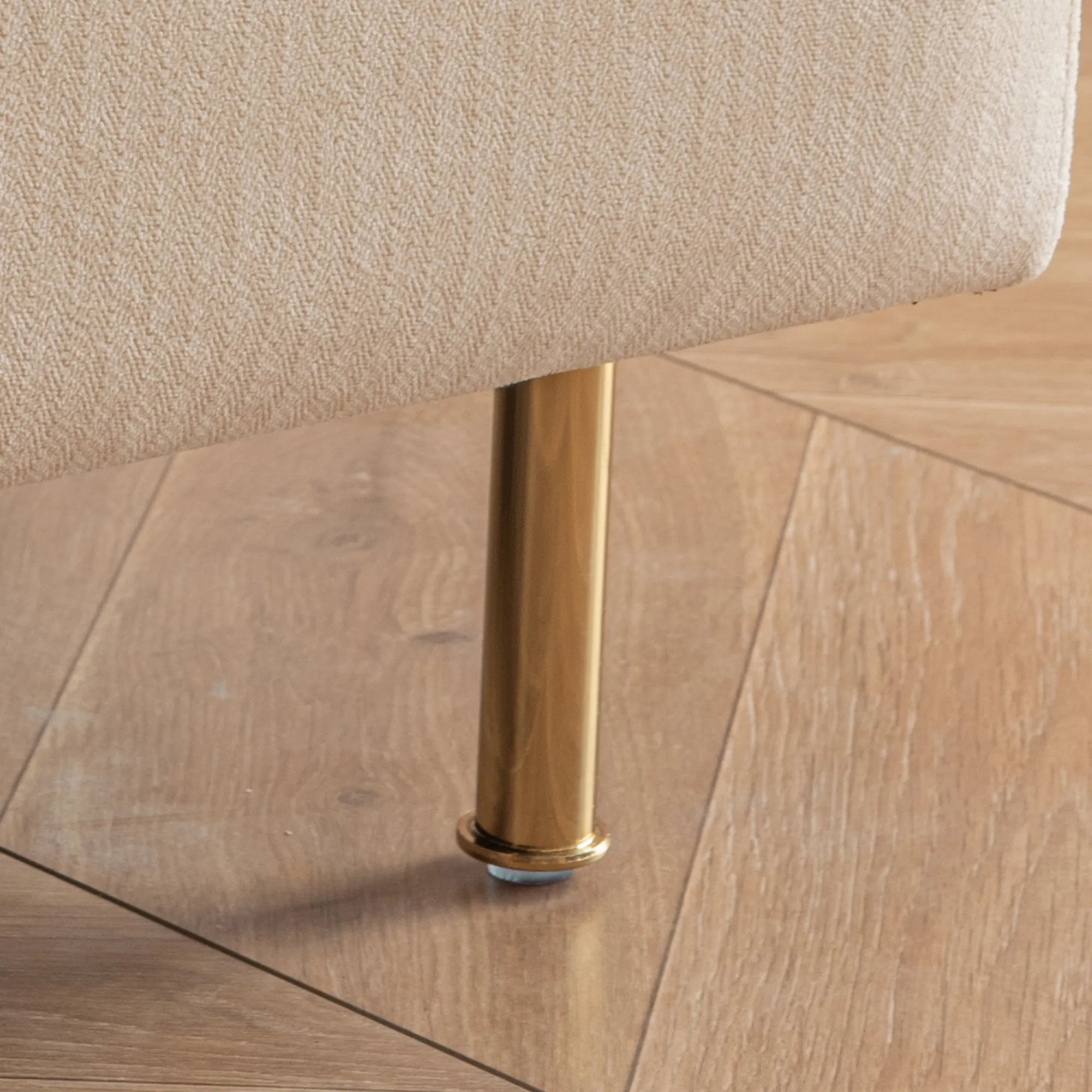 Close-up of a furniture leg in a gold color, attached to a beige upholstered MINIMALIST ACCENT CHAIR by House of Effy, standing on sturdy solid hardwood flooring with visible grain patterns.