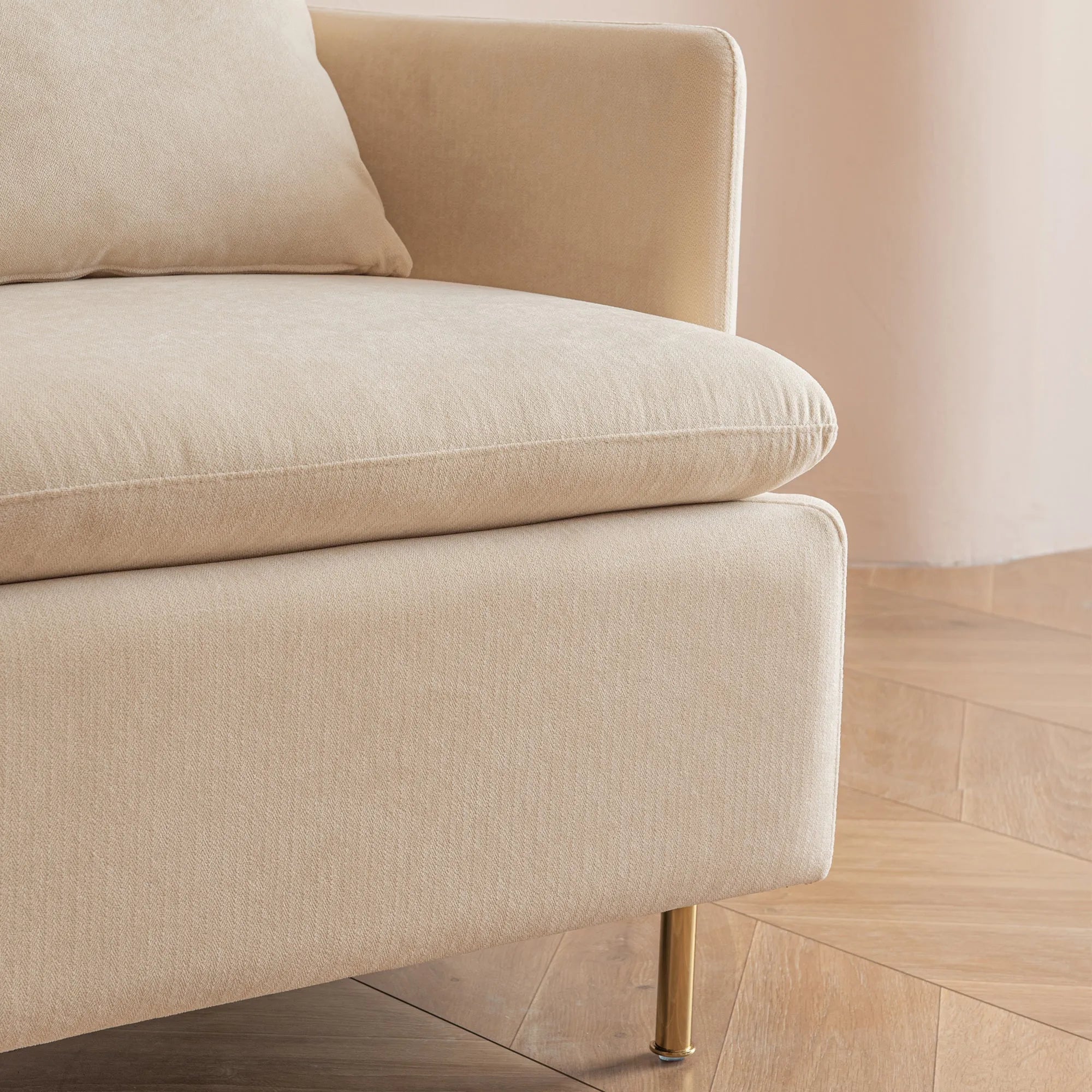 Close-up of a beige, upholstered MINIMALIST ACCENT CHAIR corner by House of Effy with a cushion, showcasing modern design with brass legs and a wooden floor in the background.