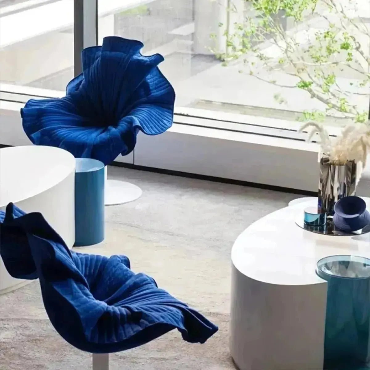 A modern room features two blue, BLOOMING CHAIR by House of Effy, a white coffee table, and a vase with dried plants near large windows overlooking greenery.