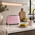 Load image into Gallery viewer, A pink retro-style SMEG toaster sits on a white marble kitchen countertop, styled with a plate of avocado toast, a white mug, a wooden cutting board, and a vase in a bright kitchen setting.
