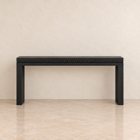 A black console table with a beaded nailhead trim along the front edge, set against a neutral beige textured wall and tiled floor.