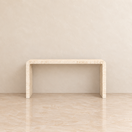 A minimalist console table with a curved top and thick blocky legs, crafted from a light beige textured stone or wood, set against a matching beige textured wall and floor.