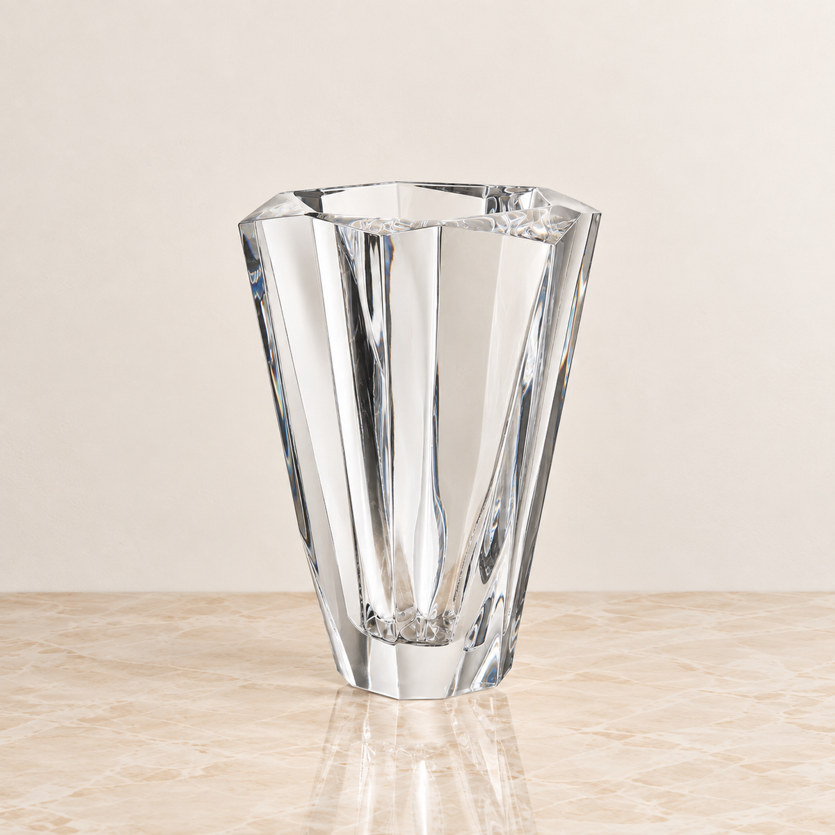 A clear faceted glass vase with a geometric, multi-sided design, standing on a light beige marble surface against a neutral cream background.