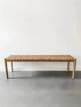 Load image into Gallery viewer, A House of Effy WOODEN CONTEMPORARY BENCH with a minimalist, sleek design and a woven brown seat is placed against a plain white wall on a gray floor. Crafted from premium quality wood, this piece embodies modern elegance.
