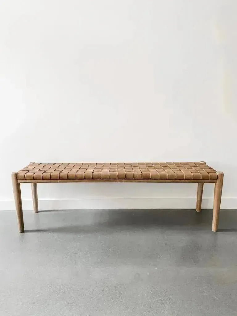 A House of Effy WOODEN CONTEMPORARY BENCH with a minimalist, sleek design and a woven brown seat is placed against a plain white wall on a gray floor. Crafted from premium quality wood, this piece embodies modern elegance.