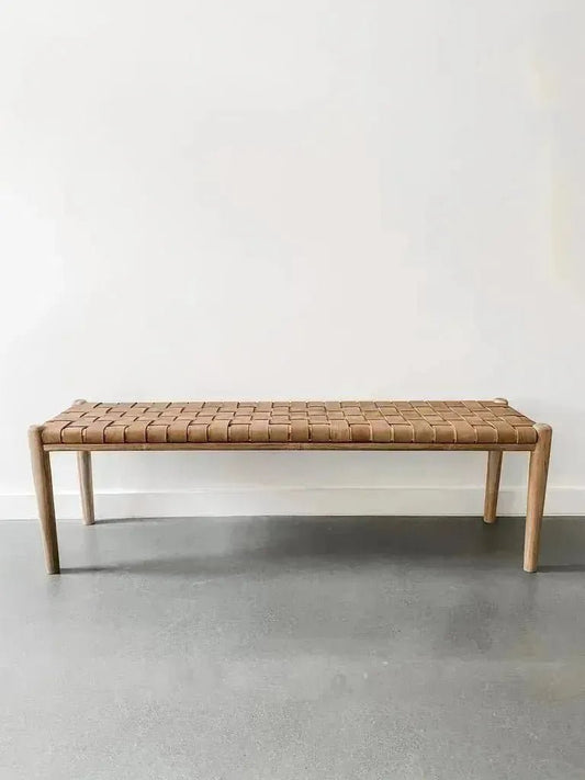 A House of Effy WOODEN CONTEMPORARY BENCH with a minimalist, sleek design and a woven brown seat is placed against a plain white wall on a gray floor. Crafted from premium quality wood, this piece embodies modern elegance.