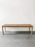 Load image into Gallery viewer, A House of Effy WOODEN CONTEMPORARY BENCH with a minimalist, sleek design and a woven brown seat is placed against a plain white wall on a gray floor. Crafted from premium quality wood, this piece embodies modern elegance.
