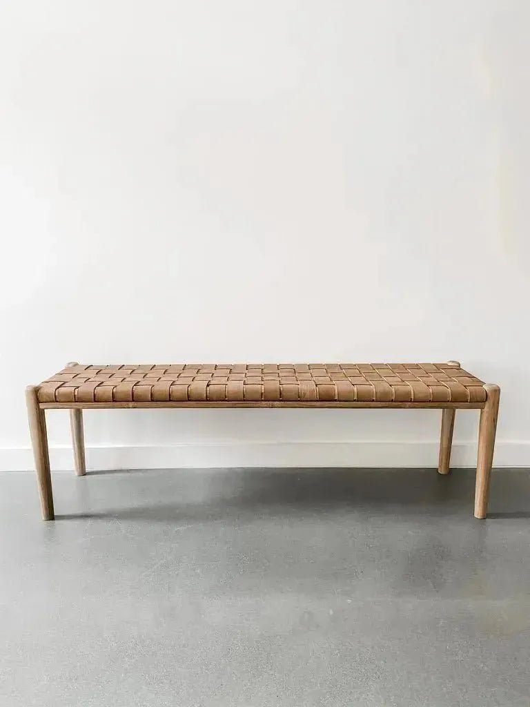 A House of Effy WOODEN CONTEMPORARY BENCH with a minimalist, sleek design and a woven brown seat is placed against a plain white wall on a gray floor. Crafted from premium quality wood, this piece embodies modern elegance.