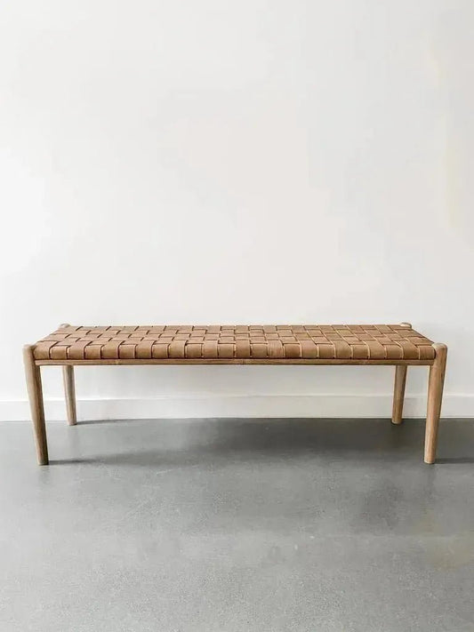 A House of Effy WOODEN CONTEMPORARY BENCH with a minimalist, sleek design and a woven brown seat is placed against a plain white wall on a gray floor. Crafted from premium quality wood, this piece embodies modern elegance.