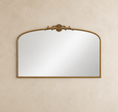 Load image into Gallery viewer, An ornate arched wall mirror with a gold-toned frame and decorative crest at the top, mounted on a textured beige wall.

