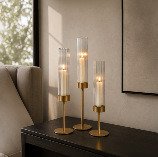 GOLD CANDLE HOLDER 3 PC IN A ROOM ON A TABLE BY ANOTHER PIECE OF FURNITURE AND WINDOW