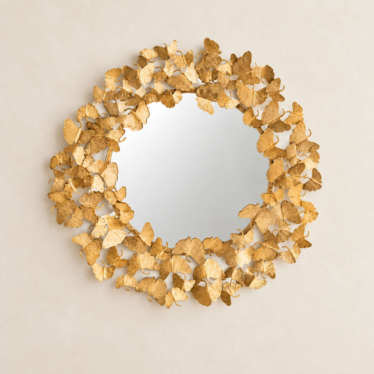 A round decorative mirror with a textured frame made of overlapping metallic gold leaf pieces, shown against a plain light beige background.