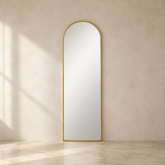 An arched full-length floor mirror with a slim gold frame, standing against a textured beige plaster wall on a light stone floor with soft window shadows.