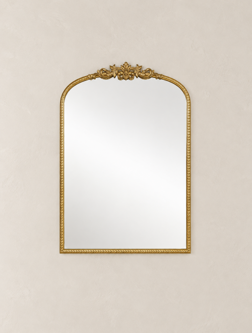 An ornate gold-framed wall mirror with a decorative crest at the top, shown against a textured beige wall.