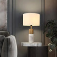 Load image into Gallery viewer, A House of Effy MODERN BEDSIDE LAMP with a white cylindrical shade and a gold and marble base stands on a round marble side table next to a gray upholstered chair and a plant, providing ambient lighting with its minimalistic design.
