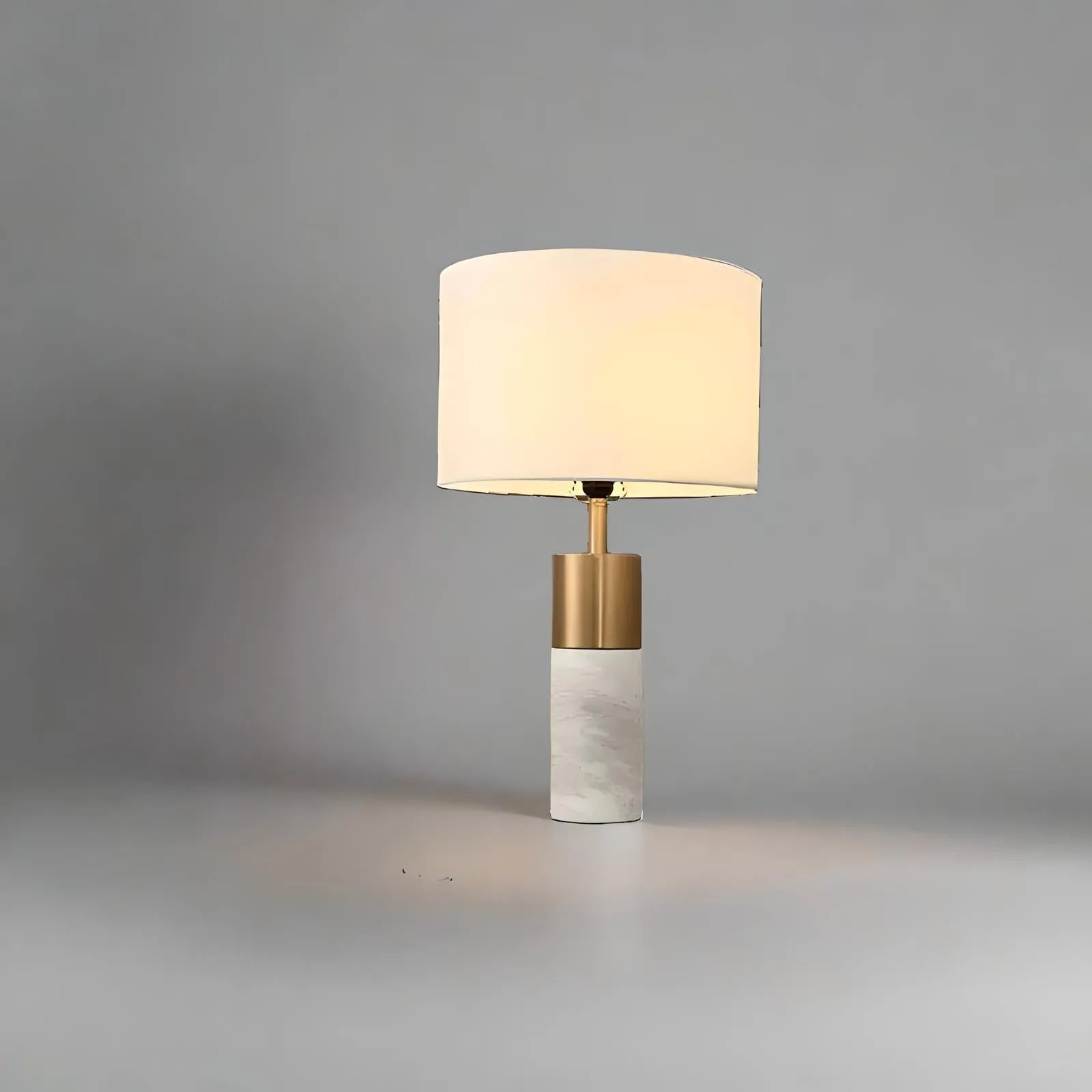 A House of Effy MODERN BEDSIDE LAMP with a cylindrical, marble-like base and a golden stem, topped with a white fabric lampshade, set against a plain grey background. This minimalistic design provides ambient lighting perfect for any contemporary bedroom.