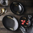 Load image into Gallery viewer, A luxurious dining experience featuring black dinnerware, including plates, bowls, and utensils. A MATTE BLACK CHARGER PLATE by House of Effy enhances the elegance, accompanied by a glass and three strawberries on a napkin.
