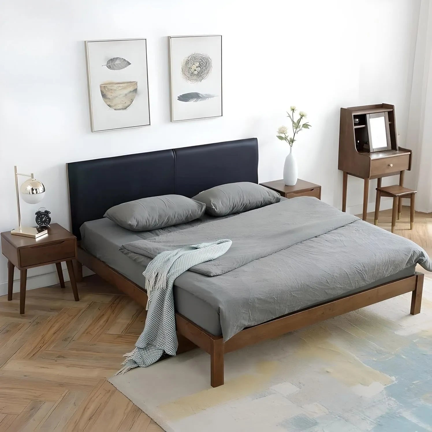 A neatly made bed with gray bedding, two pillows, and a throw blanket is positioned against a wall in this sophisticated bedroom. The House of Effy BROWN WOOD BED is flanked by nightstands with lamps and vases, while a small desk and two framed artworks adorn the wall above.