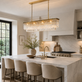 Load image into Gallery viewer, a linear crystal chandelier with hanging clear crystal prisms is illuminated above a marble kitchen island with four upholstered bar stools in a bright, modern kitchen.
