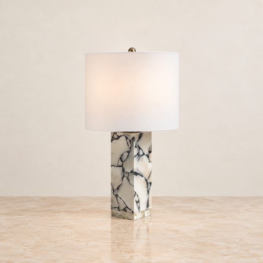 A table lamp with a white drum shade and a polished white marble base featuring black veining, set on a light beige stone surface against a neutral beige wall.