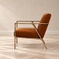 Load image into Gallery viewer, A modern accent chair with a warm rust-orange velvet upholstery and a polished gold-finished metal frame, shown in a side profile against a neutral beige wall and marble floor.

